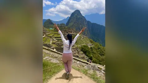 Happy traveler in white shirt and pink pants raising arms at the Machu Picchu ruins under a blue sky.