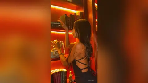 Elegant woman in a black backless dress browsing books in a warm, moody library setting.