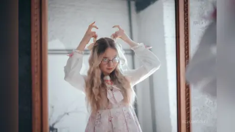 Young girl with long blonde hair wearing glasses and a pink floral dress looking in a mirror with a dreamy mood.