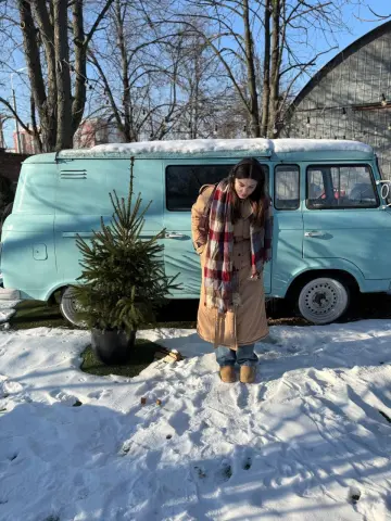 Woman in a tan coat and plaid scarf standing by a vintage light blue van in a snowy winter landscape.