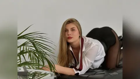 Blonde woman in white shirt and black skirt posing elegantly next to a green plant in a bright room.
