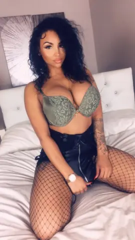 Curvy woman in a green lace bra and black leather skirt sitting on a white bed in a cozy bedroom setting.