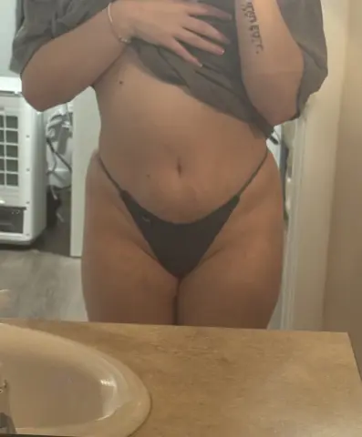 RaelynnWhitee poses in a grey thong and a grey shirt pulled up to show her stomach, showing a tattoo on her arm in a bathroom setting.