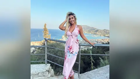 Anita stands on a balcony overlooking the ocean wearing a pink floral sundress.