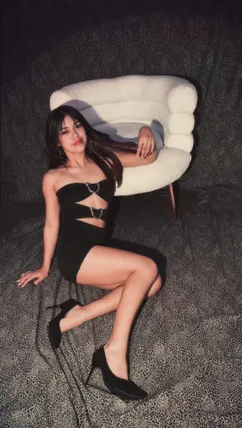 Elegant woman in a black cutout mini dress and heels sitting on a white chair against a dark textured background.