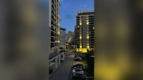 Modern high-rise apartment buildings illuminated at twilight overlooking a quiet urban street with parked cars.