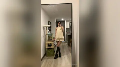 Young woman in a cream halter dress and black boots posing in a modern minimalist apartment mirror.