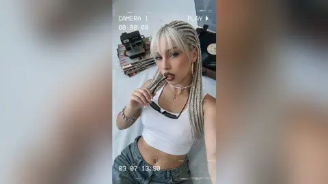 Young woman with blonde braids in a white tank top posing for a selfie in a cozy indoor setting.