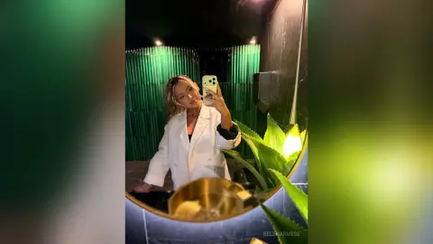 Blonde woman in a white oversized blazer taking a mirror selfie in a modern luxury bathroom with green accents.
