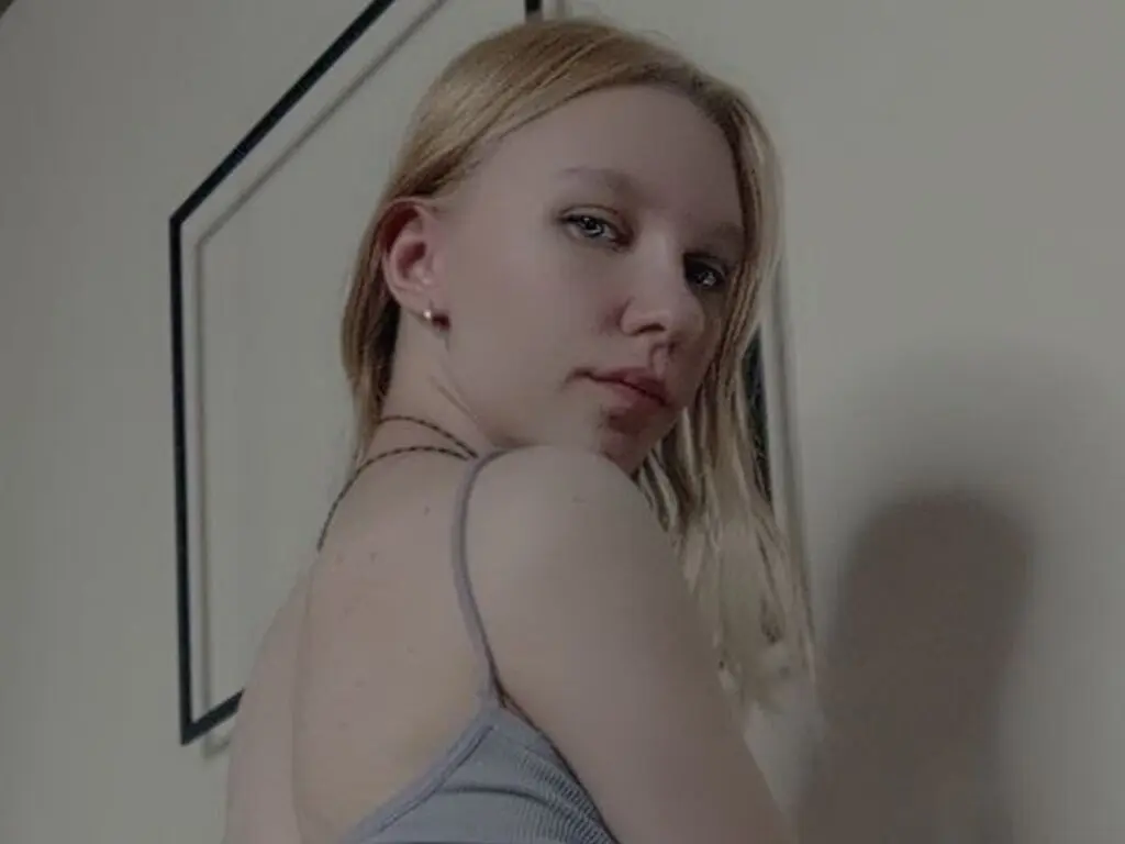 Blonde woman in a grey ribbed tank top looking thoughtfully at the camera in a minimalist indoor setting.
