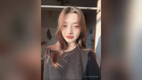 Young woman with long wavy brown hair wearing a cozy black ribbed sweater in warm sunlight.