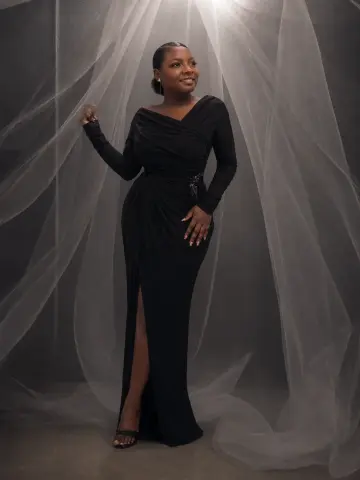 LilyReeds smiles in a long black evening gown with a high leg slit, posing against a backdrop of sheer white fabric.