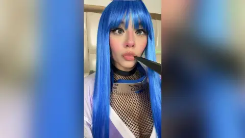 Cosplayer with long blue hair wearing a black mesh outfit in a soft indoor setting.