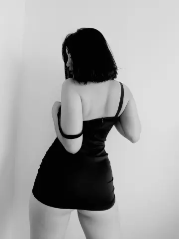 AnnaKomatsuu poses from behind wearing a tight black mini dress that shows her bare back and legs.