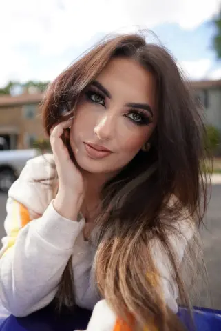 Beautiful woman with long brown hair wearing a white sweatshirt in a bright outdoor setting.