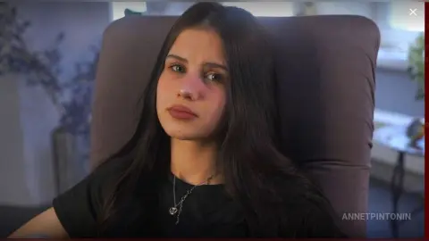 Young woman with long dark hair wearing a black t-shirt and silver necklace in a moody indoor setting.