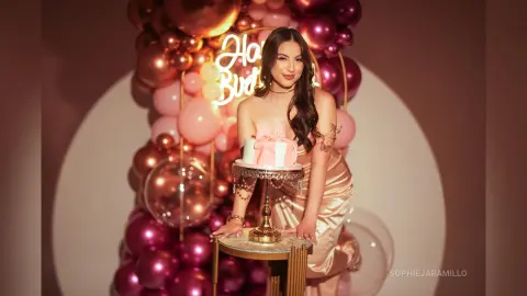 Elegant woman in a champagne satin dress celebrating her birthday with a cake and pink balloon decor.