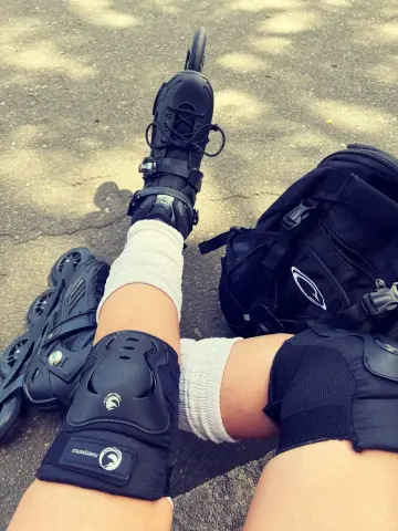 Person in knee pads and white socks sitting on pavement with black inline skates and a backpack on a sunny day.