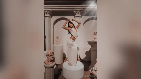 Elegant woman in a white backless gown posing amidst classical marble statues in a sunlit museum setting.