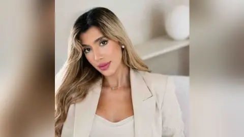 Elegant woman in a beige blazer and necklace posing with a sophisticated mood in a bright minimalist room.
