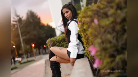 Stylish woman in a white shirt and black shorts sitting on a stone ledge near garden bushes at sunset.
