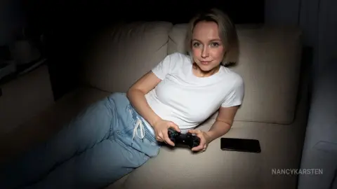 NancyKarsten — Young blonde woman in white t-shirt and blue sweatpants relaxing on a sofa with a gaming controller.