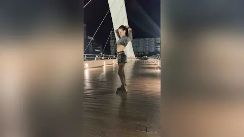 Stylish woman in a grey crop top and denim shorts posing on a city boardwalk at night.