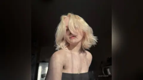 Blonde woman with wavy hair in a dark room, wearing a black strapless top with a moody, cinematic lighting vibe.