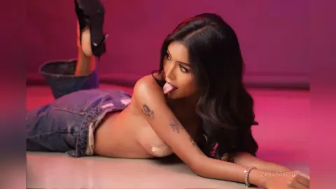 Young woman in denim jeans lying on a pink floor with a playful expression and long dark hair.
