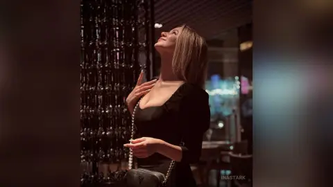 InaStark poses in a black dress with her head tilted back and eyes closed in a dimly lit setting.