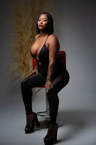 Confident Black woman in a sleek black bodysuit sitting on a red chair against a neutral studio backdrop.