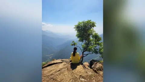 Woman in a yellow shirt sitting on a mountain peak overlooking a misty valley landscape.