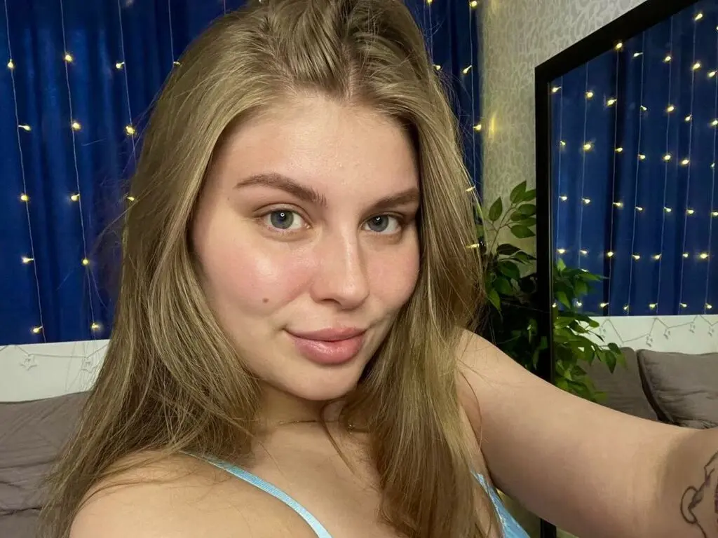 Smiling blonde woman in a light blue camisole relaxing in a cozy bedroom with fairy lights.