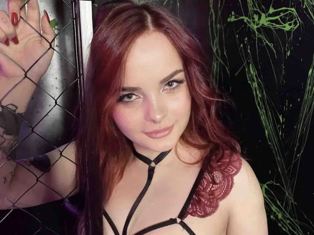 Smiling woman with red hair in burgundy lace lingerie posing near a chain-link fence in a moody setting.