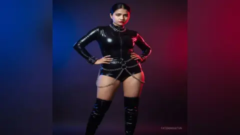 Young woman in a sleek black latex bodysuit and tall boots posing against a moody blue and red studio background.