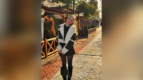 Blonde woman in a black dress and white sherpa jacket standing on a sunny cobblestone street.