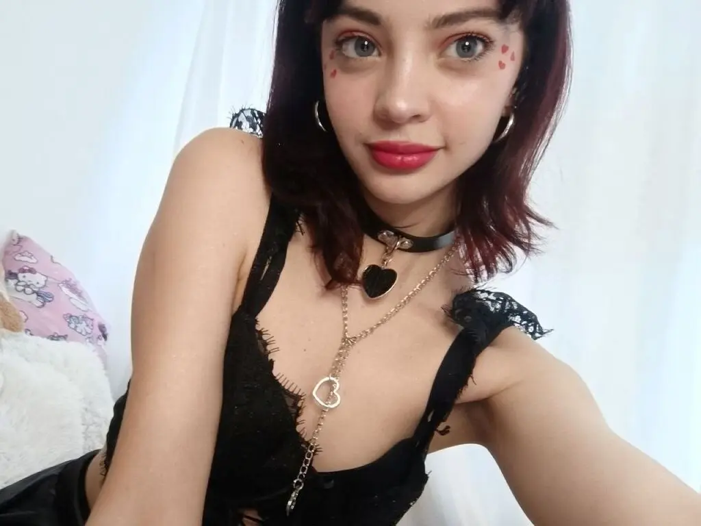 Young woman with dark hair wearing a black lace top and heart necklaces in a bright indoor setting.