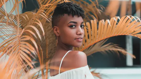 AnnPrice poses looking over her shoulder, wearing a white off-the-shoulder top with her short dark hair and gold hoop earrings, framed by large orange palm leaves.