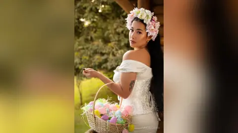 JuliahLewis poses outdoors wearing a white off-the-shoulder lace corset and a pink and white flower crown.