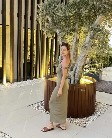 Woman in an olive green maxi dress standing by a large tree in a modern outdoor patio setting.