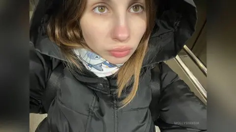 Young woman in a black hooded puffer jacket and patterned scarf looking thoughtfully at the camera.