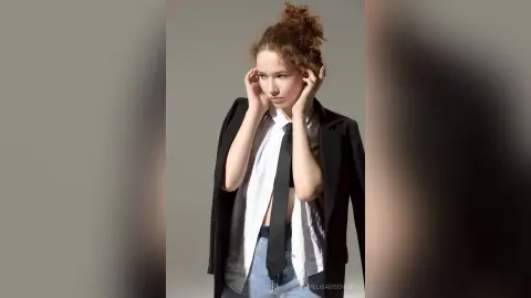 Young woman in a white shirt, black tie, and blazer posing with a thoughtful expression against a grey background.
