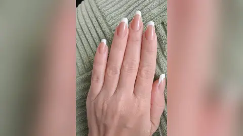 Elegant nude French manicure with white tips on long almond shaped nails against a soft sage green fabric.