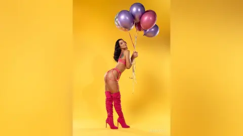 Glamorous woman in pink lingerie and tall boots holding colorful balloons against a bright yellow background.