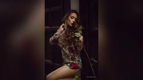 Elegant woman in a floral print dress holding a red rose in a moody, dark indoor setting.
