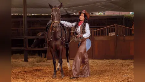 Cowgirl in western attire leaning against a brown horse in a rustic ranch stable setting.
