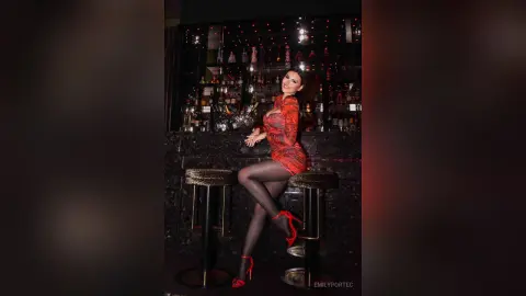 Elegant woman in a red patterned dress and heels sitting on a bar stool in a moody, dimly lit lounge.