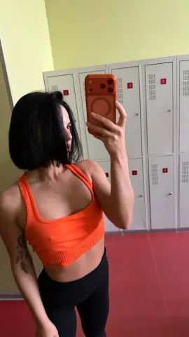 Woman in orange sports bra and black leggings taking a mirror selfie in a gym locker room.