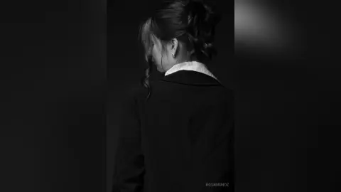 Moody black and white portrait of a woman in a dark blazer and white shirt looking away in a dim setting.