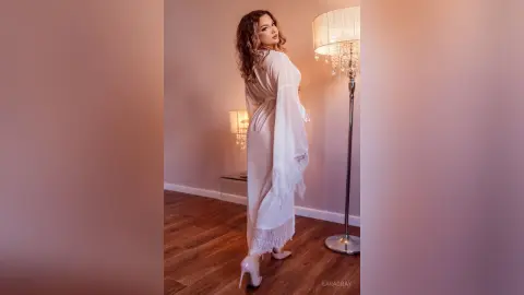 Elegant woman in a white silk robe standing in a warm, dimly lit room near a crystal floor lamp.